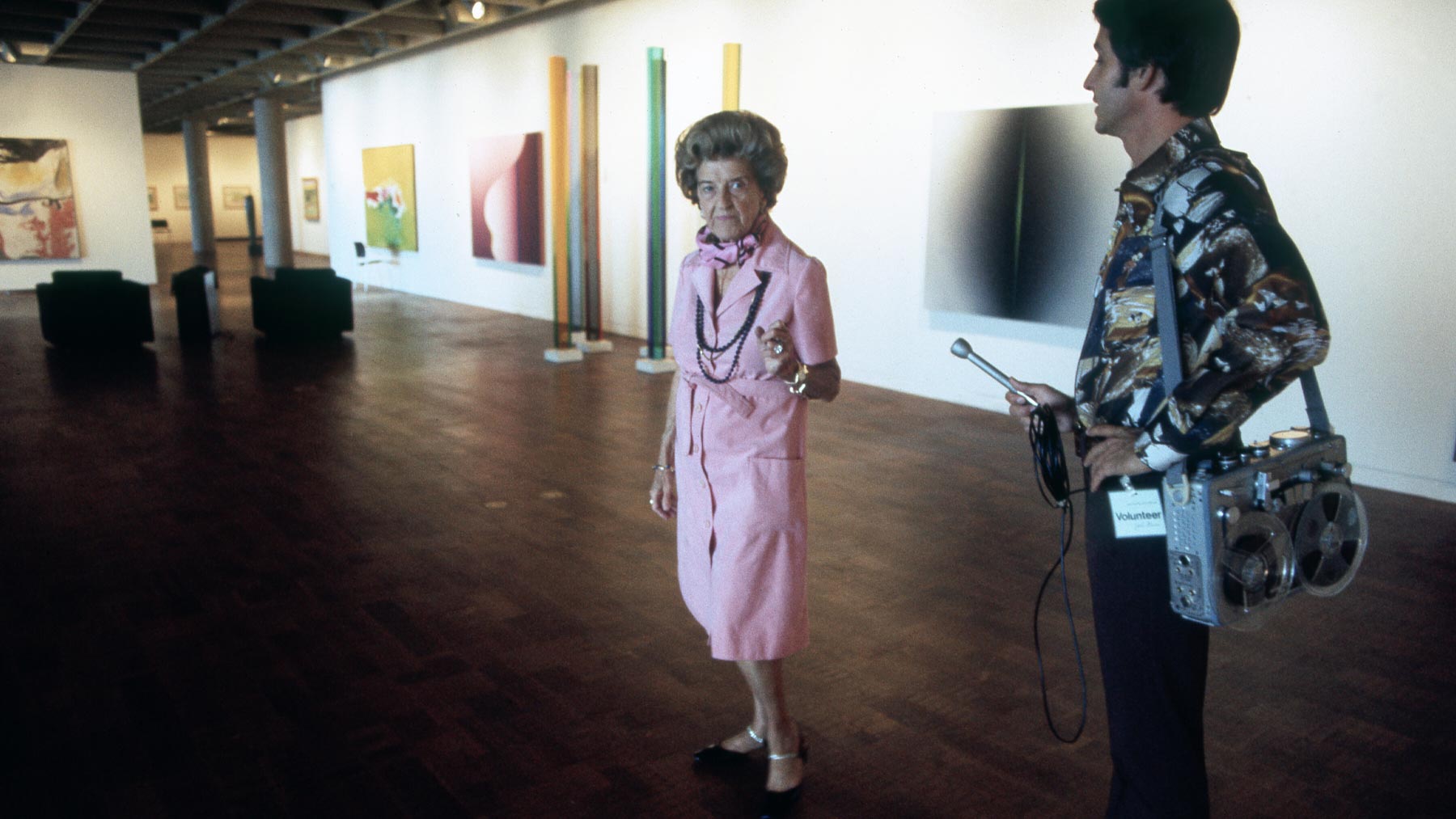 A woman in a pink outfit stands in an art gallery, engaging with a man holding a microphone and recording device. Colorful abstract artworks are displayed on the walls.